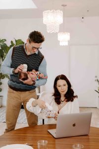 Father holding baby while feeding bottle of milk while mom looks at computer